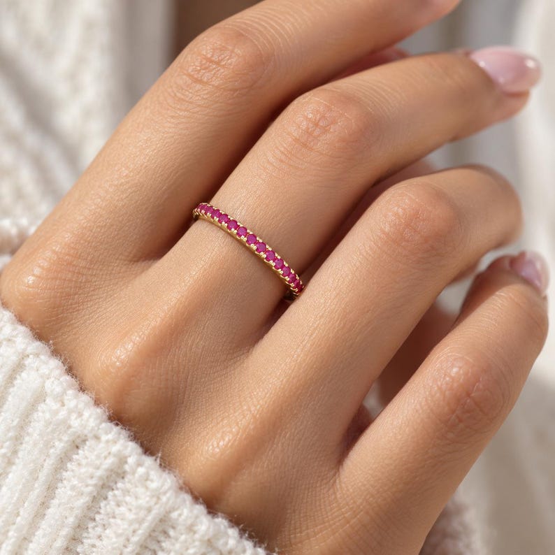 May include: A thin gold band ring featuring a continuous row of small, round, vibrant pink gemstones set along the entire outer circumference. The metal has a polished finish, and the stones are uniform in size, creating a delicate, circular design.