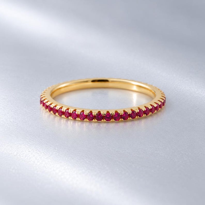 May include: A thin gold band ring featuring a continuous row of small, round, vibrant red gemstones set along the outer circumference. The metal has a polished, reflective finish, and the stones are evenly spaced in a classic eternity style design.