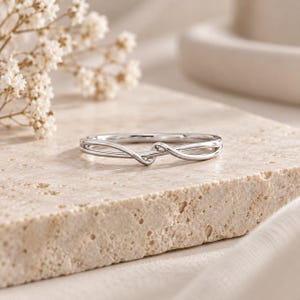 Può includere: Un anello color argento lucido caratterizzato da una fascia sottile e delicata che si curva e si sovrappone al centro per creare un design sottile e continuo simile a un nodo. La superficie metallica è liscia e riflettente e pulita.