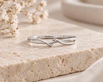 May include: A polished silver-toned ring featuring a delicate, slender band that curves and overlaps at the centre to create a subtle, continuous knot-like design. The metal surface is smooth and reflective, showcasing a clean, minimalist aesthetic.