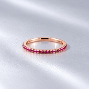 May include: A rose gold eternity band featuring a continuous row of small, round, vibrant red gemstones set along the entire circumference of the thin metal ring, creating a uniform and polished appearance with a bright, reflective finish.