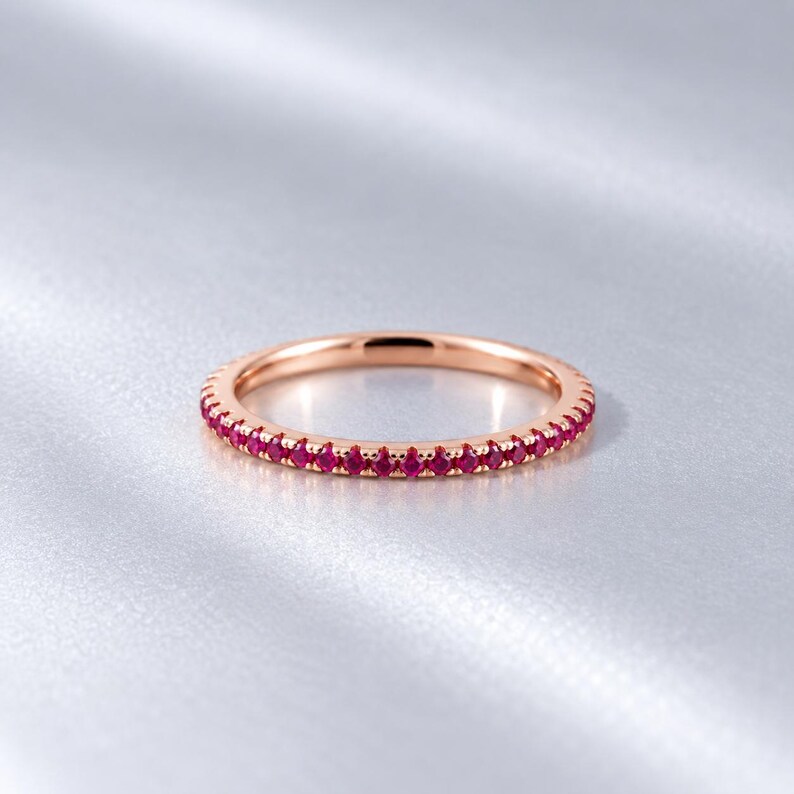 May include: A rose gold eternity band featuring a continuous row of small, round, vibrant red gemstones set along the entire circumference of the thin metal ring, creating a uniform and polished appearance with a bright, reflective finish.