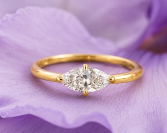 May include: A gold ring featuring a single marquise-cut clear gemstone set in a four-prong setting. The band is smooth and polished, showcasing a minimalist design that highlights the elongated shape of the central stone against a purple background.