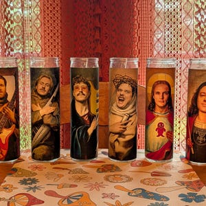 May include: Six cylindrical glass prayer candles, each featuring a unique portrait of a person in a religious-style robe. The designs include individuals holding a bass guitar, a keytar, and one depicted with a crown of thorns and a red heart emblem.
