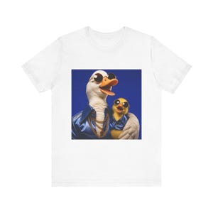 May include: A white t-shirt featuring a graphic print of an adult duck and a duckling wearing blue jackets, gold chains, and round sunglasses. The adult duck has white feathers and the duckling is yellow, both set against a solid blue background.