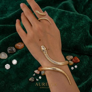May include: A gold snake-shaped bracelet that wraps around the wrist and a matching gold snake-shaped ring on the finger. The jewelry features a textured, scale-like pattern. The text AUREVIA JEWELLERY is printed at the bottom of the image.