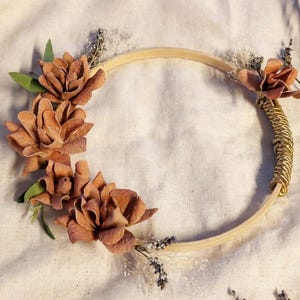 May include: A circular wooden embroidery hoop decorated with brown dried flowers, green leaves, and small sprigs of white baby's breath. One side of the hoop is wrapped in gold-colored wire, and additional dried lavender stems are attached.