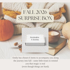 May include: A white rectangular box with the text "Contains 6 items" printed on the front. The box is centered against a background featuring a stack of folded knit sweaters, a pumpkin, a candle in a wooden bowl, and a book.