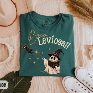 May include: A dark teal t-shirt featuring a graphic of a ghost wearing a witch hat, holding an open book and a magic wand. A black cat floats nearby. The text "Booo Leviosa!!" is printed in a cursive and serif font above the ghost illustration.