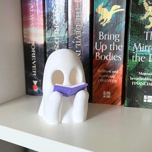 May include: A white, ghost-shaped figurine with two hollow eye sockets, holding a small, open purple book with white pages. The ghost has a smooth, rounded head and two small arms positioned in front to support the book, standing upright.