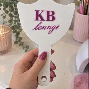 May include: A white handheld mirror with a shield-shaped head and a handle featuring a circular cutout at the base. The surface displays the text KB lounge in a dark pink, raised, serif and script font combination centered on the mirror face.