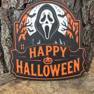 May include: A layered wall sign featuring a black ghost figure in front of a white full moon, orange trees, and a haunted house silhouette. The text HAPPY HALLOWEEN is displayed in orange letters above a small orange jack-o'-lantern icon.