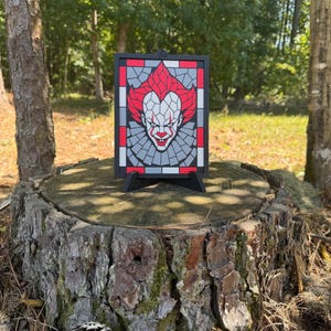 May include: A rectangular black-framed wall art piece featuring a mosaic-style graphic of a clown with red hair, a white face, and a red smile. The design uses geometric segments in red, white, gray, and black, displayed on a black plastic stand.