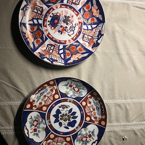 May include: Two round ceramic plates featuring intricate patterns in deep blue, vibrant orange, and white. The top plate displays floral motifs and architectural elements, while the bottom plate showcases a central flower surrounded by panels of birds and blossoms.