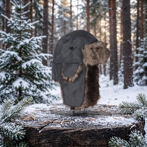 May include: A charcoal gray trapper hat featuring a smooth, water-resistant outer shell and thick, brown faux fur lining. The hat includes ear flaps that can be secured with a small black plastic buckle strap at the bottom.