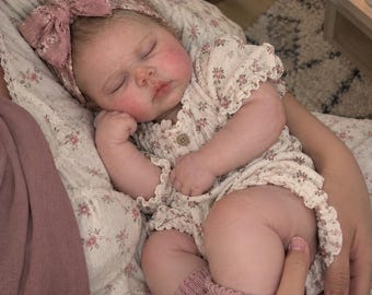 May include: A realistic baby doll with closed eyes and flushed cheeks, wearing a cream-coloured romper with a small floral pattern, a matching dusty pink headband with a large bow, and knitted dusty pink socks with small bows on the ankles.