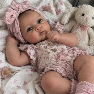 May include: A realistic baby doll with dark eyes and rosy cheeks, wearing a pink floral romper with ruffled sleeves, two wooden buttons, and a matching bow headband. The doll is paired with pink knit socks, a light pink pacifier, and a plush white bunny.