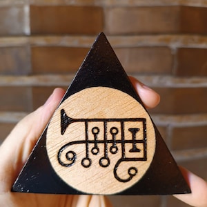 Op de afbeelding: Een driehoekig houten object met een zwart geschilderde rand en een centrum van natuurlijk hout. Het midden bevat een donker, ingebrand zegelontwerp bestaande uit een trompetvorm, meerdere cirkels en lijnen die een complex symbool vormen.