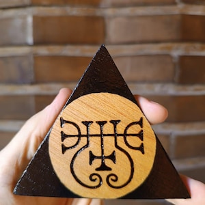 May include: A triangular wooden object with a dark stained border and a natural light wood centre. A black circular design featuring a complex, symmetrical sigil with cross and scroll elements is burned into the centre of the light wood surface.