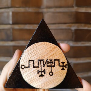 Op de afbeelding: Een driehoekig houten object met een zwart geschilderde rand en een cirkelvormig midden van natuurlijk licht hout. Het midden bevat een donker, ingebrand lineair symbool met kruisachtige elementen en cirkelvormige uiteinden.