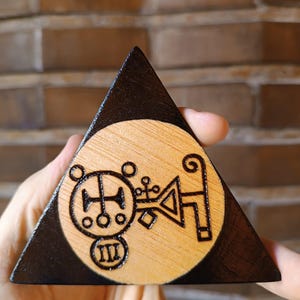 Könnte beinhalten: Eine dreieckige Holzplatte mit einem dunkel gebeizten Rand und einer Mitte aus hellem Naturholz. Die Mitte zeigt ein schwarzes, in das Holz eingebranntes Siegeldesign mit kreisförmigen Elementen, geometrischen Linien und der römischen Ziffer III.