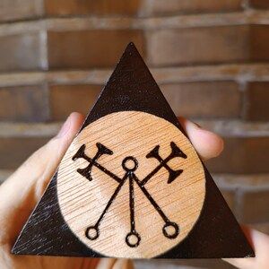 Peut inclure: Une pièce triangulaire en bois avec un centre circulaire en bois naturel et une bordure en bois foncé. Le centre présente un motif pyrogravé de deux haches croisées au-dessus de trois petits cercles reliés par des lignes, formant un motif géométrique.