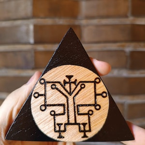 Peut inclure: Une plaque triangulaire en bois avec une bordure peinte en noir et une incrustation circulaire en bois naturel au centre. L'incrustation présente un sceau géométrique sombre, complexe et symétrique, gravé avec des lignes fines et précises.