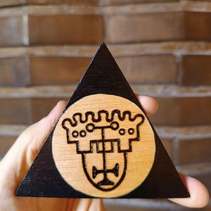 May include: A triangular wooden plaque with a black painted border and a natural wood centre. The centre features a dark, burned-in wood engraving of a stylised crown symbol with a vertical line and circular details, creating a distinct contrast.