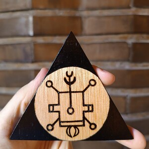 Op de afbeelding: Een driehoekig houten object met een zwart geschilderde rand en een centrum van natuurlijk hout. Het midden bevat een cirkelvormig in het hout gebrand ontwerp bestaande uit een centrale rechthoek, lijnen, cirkels en halve manen.