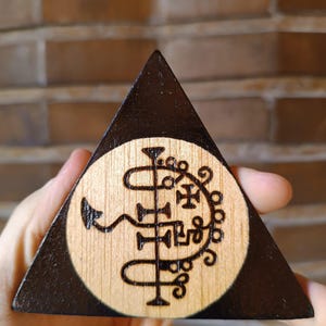 Pode incluir: Uma peça triangular de madeira com uma borda tingida de escuro e um centro de madeira clara natural. O centro apresenta um design circular pirografado composto por linhas pretas intrincadas, laços, uma pequena cruz e uma forma de cauda.