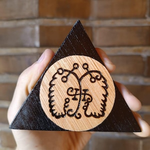Op de afbeelding: Een driehoekig houten object met een zwart gebeitste rand en een cirkelvormig lichtgekleurd houten midden. Het midden bevat een donker, ingewikkeld en symmetrisch brandmerkontwerp bestaande uit twee gespiegelde, bladachtige patronen.
