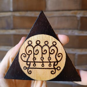Pode incluir: Uma placa triangular de madeira com um centro circular em acabamento de madeira clara natural e um design de selo queimado escuro. A moldura triangular circundante é pintada de preto fosco sólido, criando um contraste com o centro.