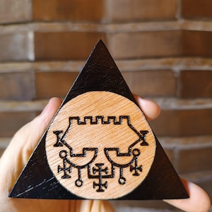 Könnte beinhalten: Eine dreieckige Holzplakette mit schwarz lackiertem Rand und einer Mitte aus Naturholz. Die Mitte zeigt ein kreisförmiges, eingebranntes Design, das ein symmetrisches, komplexes Siegel mit gezacktem Oberteil und kreuzartigen Symbolen darstellt.