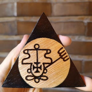 Op de afbeelding: Een driehoekige houten plaquette met een donkerzwart gebeitste rand en een natuurlijk licht houten cirkelvormig midden. Het midden toont een donker, ingewikkeld, ingebrand zegelontwerp met een kruisachtig symbool en een drietandige uitbreiding.