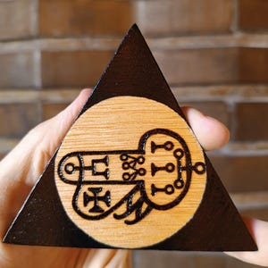 Könnte beinhalten: Eine dreieckige Holzplakette mit einem dunkel gebeizten Rand und einer Mitte aus hellem Naturholz. Ein schwarzes, in das helle Holz eingebranntes Siegeldesign ist mittig platziert und besteht aus miteinander verbundenen Linien, Kreisen und einem Kreuz.