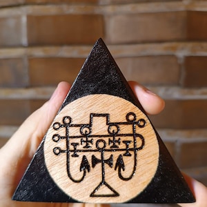 Peut inclure: Un objet triangulaire en bois avec une bordure peinte en noir et un centre en bois naturel. Le centre présente un motif de sceau circulaire sombre gravé au feu, composé de lignes horizontales, de barres verticales, de cercles et de deux crochets.
