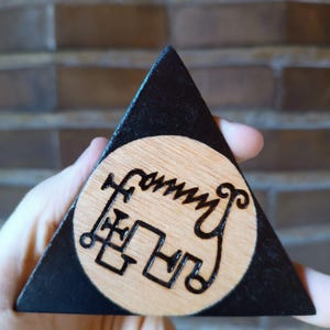 Op de afbeelding: Een driehoekig houten object met een zwart geschilderde rand en een centrum van natuurlijk hout. Het midden bevat een donker, ingebrand zegel of symbool met ingewikkelde lijnen en cirkelvormige uiteinden. De vorm is een platte, gelijkzijdige driehoek.