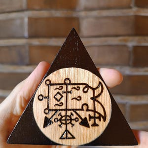 Puede incluir: Una placa triangular de madera con un borde pintado de negro y un centro de madera natural. El centro muestra un diseño circular pirograbado con intrincadas líneas geométricas negras, símbolos y un emblema central en forma de cruz.