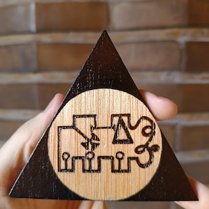 Pode incluir: Um objeto triangular de madeira com uma borda tingida de escuro e um centro circular de madeira clara natural. O centro apresenta um design gráfico preto pirografado composto por formas geométricas, linhas e um pequeno triângulo conectados.