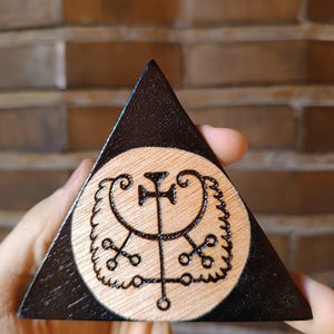 Peut inclure: Une plaque triangulaire en bois avec une bordure teintée en noir et un centre en bois clair naturel. Un sceau sombre et complexe est gravé au centre circulaire, montrant une ligne verticale avec une traverse et des volutes symétriques.