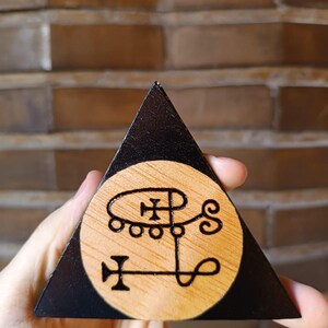 May include: A triangular wooden object with a black painted border and a natural light wood circular centre. The centre features a dark, intricate, hand-burned sigil design consisting of interconnected lines, circles, and a cross-like symbol.
