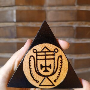 May include: A triangular wooden plaque featuring a dark stained border and a natural wood centre. The centre displays a circular wood-burned design of a complex geometric sigil with a central cross, flanked by two curved, textured wing-like shapes.