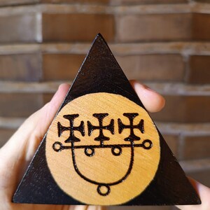 Puede incluir: Un objeto triangular de madera con un borde pintado de negro y un centro de madera clara natural. El centro presenta un diseño circular pirograbado que consiste en tres símbolos de cruz conectados por una línea curva con círculos en los extremos.