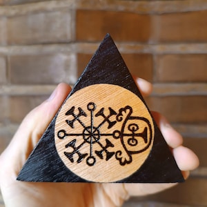 Op de afbeelding: Een driehoekig houten object met een zwart geschilderde rand en een centrum van natuurlijk hout. Het midden bevat een cirkelvormig in het hout gebrand ontwerp met een complex geometrisch symbool met stralende lijnen en een gestileerde figuur.