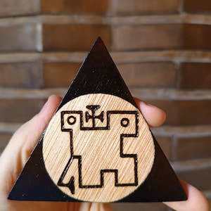 Op de afbeelding: Een driehoekige houten plaquette met een donkerzwart gebeitste rand en een cirkelvormig midden van natuurlijk licht hout. Het midden toont een donker, ingebrand geometrisch symbool dat lijkt op een gestileerd wezen met een kruisvormig embleem.