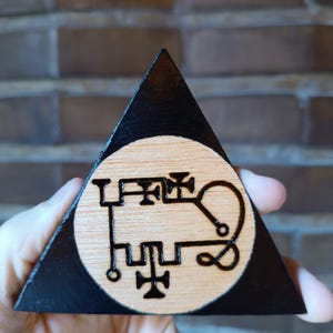 Peut inclure: Une plaque triangulaire en bois avec une bordure peinte en noir et un centre circulaire en bois clair naturel. Le centre présente un motif de sceau sombre et complexe, gravé à la main avec des lignes géométriques, des boucles et des symboles.