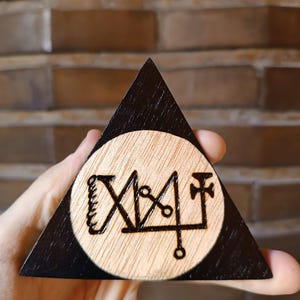 Pode incluir: Um objeto triangular de madeira com uma borda pintada de preto e um centro circular de madeira clara natural. O centro apresenta um design de selo escuro, intrincado e queimado à mão, consistindo em linhas, círculos e símbolos em forma de cruz.