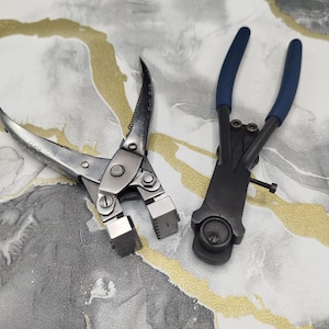May include: A pair of silver-toned metal canvas stretching pliers with a wide jaw and a pair of black metal pliers with blue rubber-coated handles and a circular head mechanism, both designed for manual gripping and tensioning tasks.