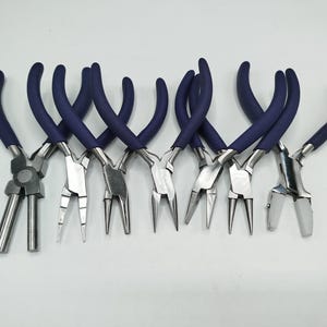 Op de afbeelding: Een set van zeven metalen sieradentangen met gepolijste zilverkleurige stalen koppen en ergonomische donkerblauwe rubberen handgrepen. De collectie bevat diverse puntvormen zoals ronde, platte, gebogen en nylon bekontwerpen voor handwerk.