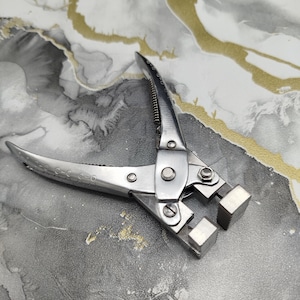 Puede incluir: Una herramienta manual de metal plateado con dos mangos curvos, un punto de pivote central con un tornillo visible y un mecanismo de resorte. La herramienta tiene dos mordazas planas, rectangulares y dentadas para sujetar o doblar.
